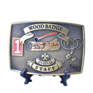 Antique brass-tone Wood Badge staff belt buckle with an axe and log motif, raised banners and colored details.