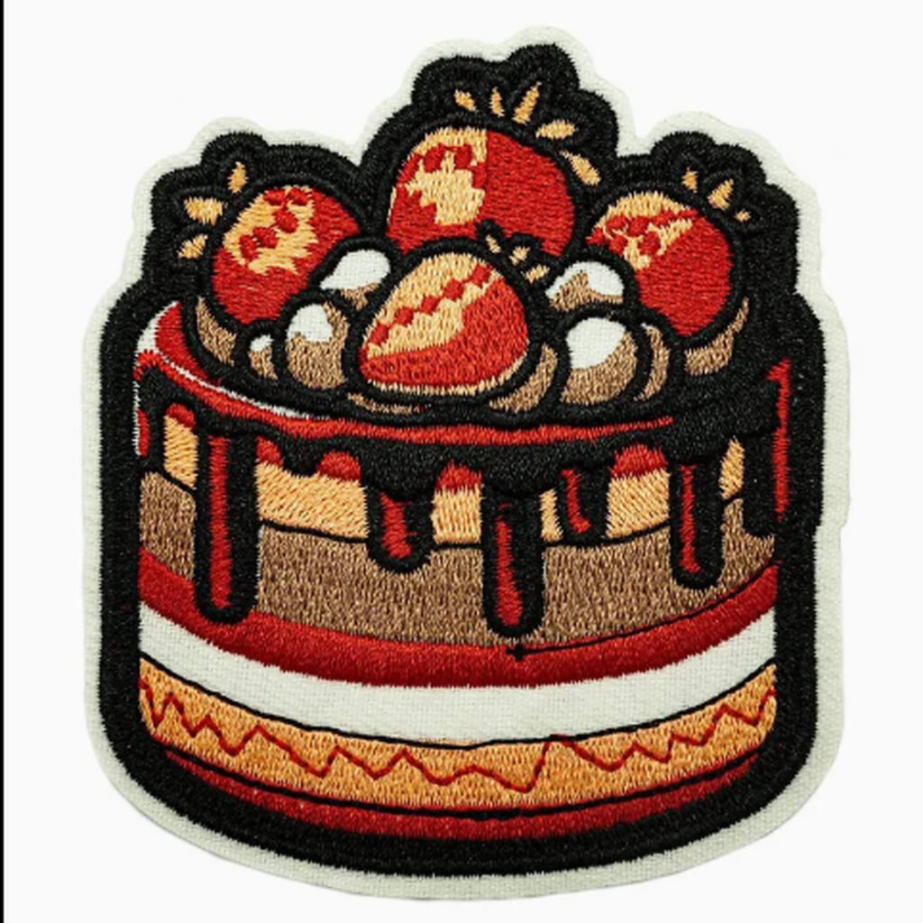 Emblema bordado em forma de bolo com morangos vermelhos, detalhes em creme, recheio em camadas e cobertura escura sobre um fundo branco.